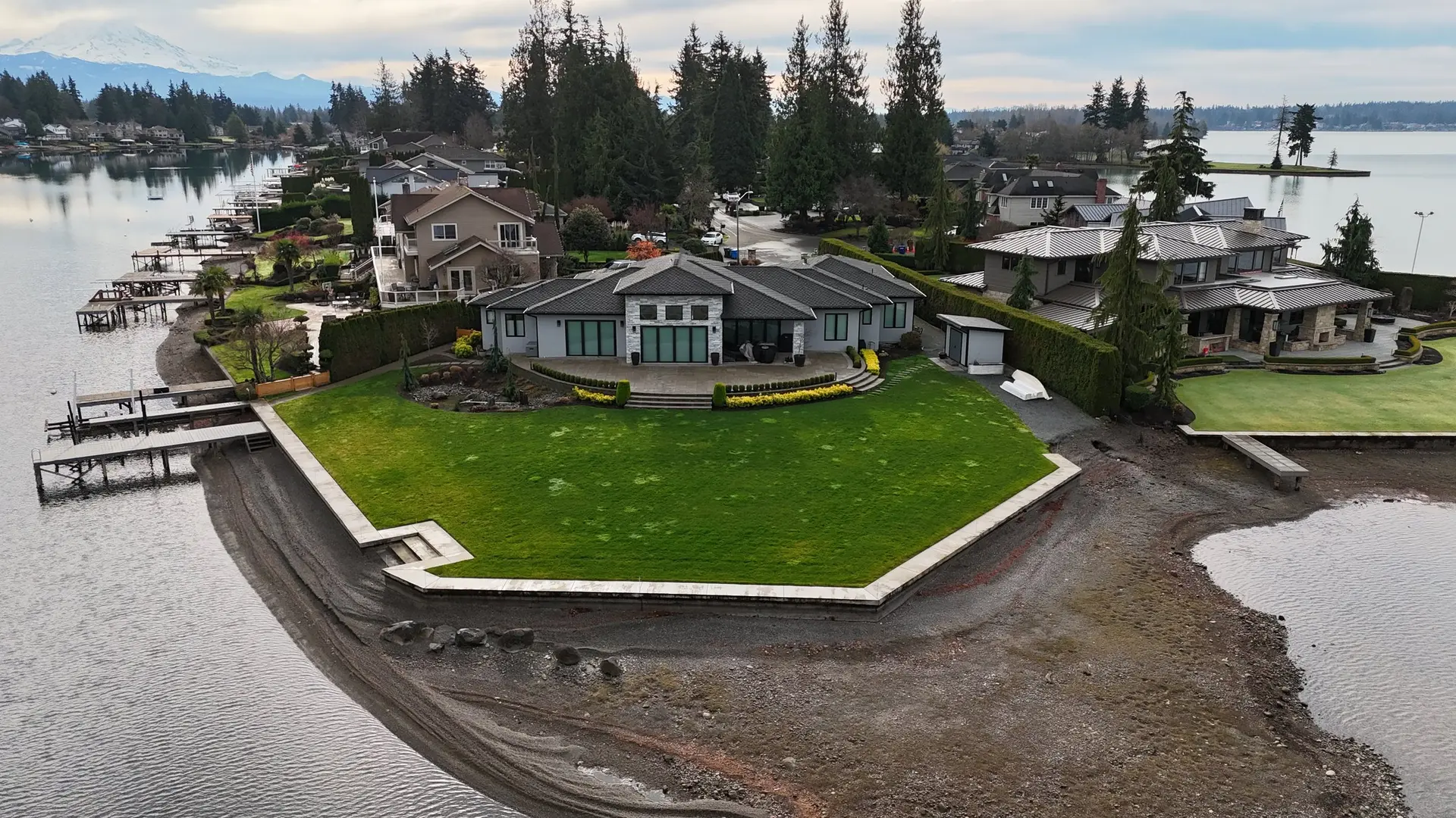 Aerial view of the finished single-storey home on a point lot, lawn running to a bulkhead on two sides, Mount Rainier on the horizon