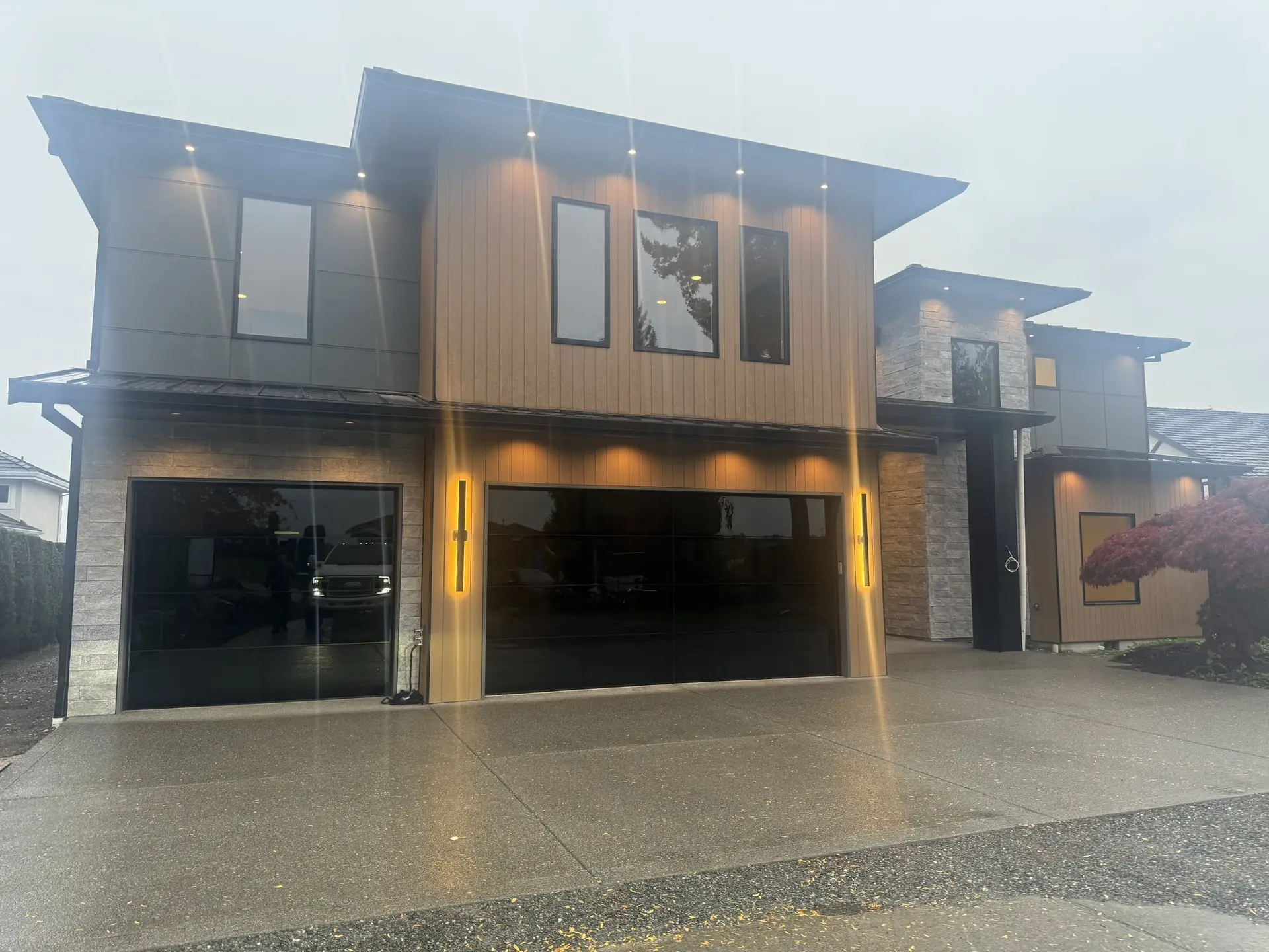 Finished street elevation at dusk in vertical cedar and stone, black glazed garage doors, soffit lighting on