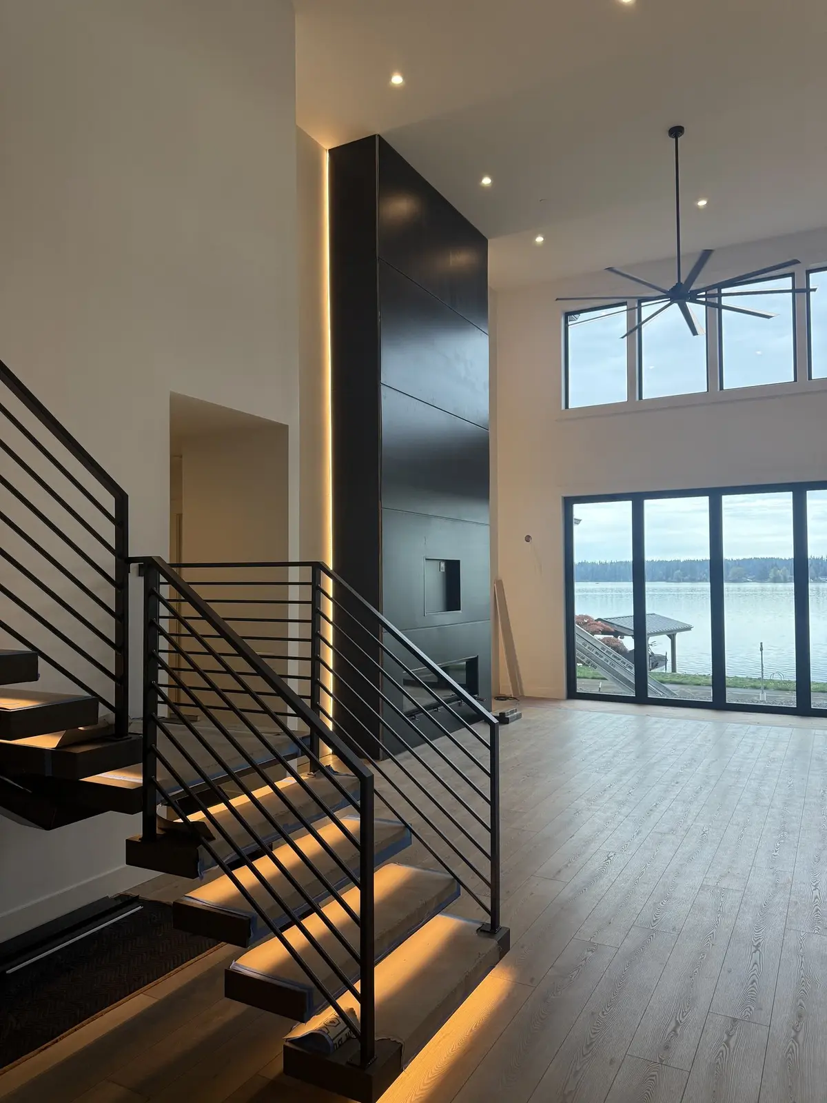 Floating stair on blackened steel stringers with lit treads, beside a full-height steel fireplace surround and windows to the lake