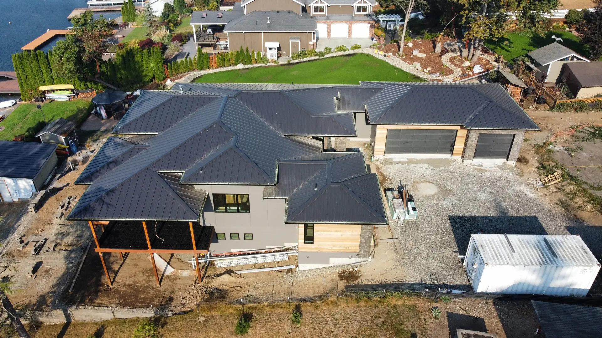 Aerial view of a lakefront home under a standing-seam metal roof, cedar and stone at the garage, dock and open water beyond